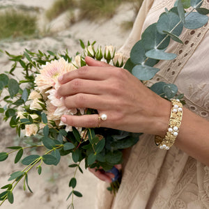 Bouquet of pink flowers and eucalyptus leaf with a hand wearing a solitaire saltwater akoya pearl ring and wide floral pearl and yellow gold bracelet. 