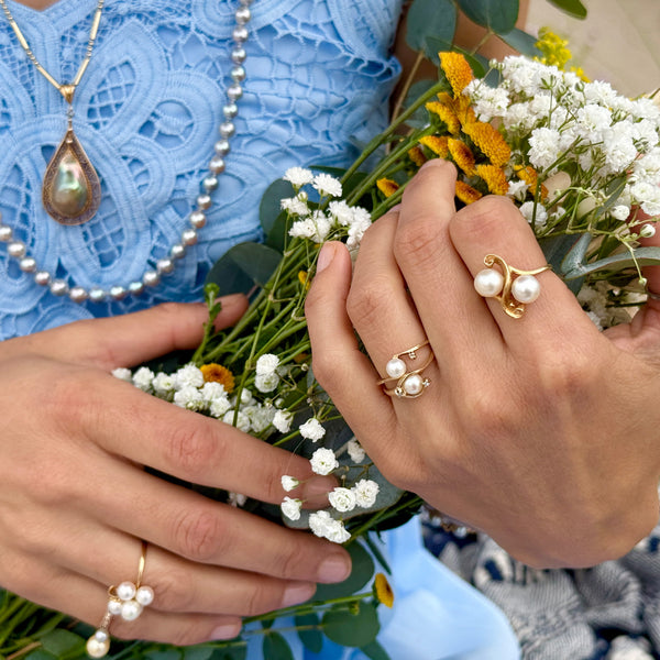 Close-up of hands wearing white akoya pearl rings with a bouquet of flowers and a blue dress.