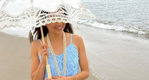 Woman on a beach wearing a blue lace dress with multiple strands of beautiful saltwater akoya pearls holding a cream lace umbrella.