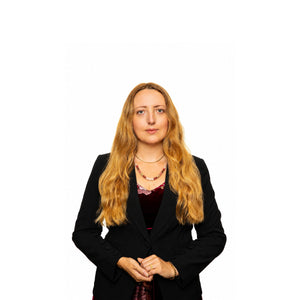 Blond woman in a black blazer on a white background wearing a shell and gold necklace.