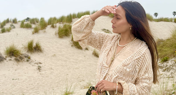 Woman in a beige lace dress standing on sandy dunes with sea grass and a white akoya pearl necklace, bracelet and earrings.