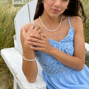 Woman in a blue lace dress sitting on a white wood chair with sea grass in the background and wearing a white pearl bracelet, ring and necklace.