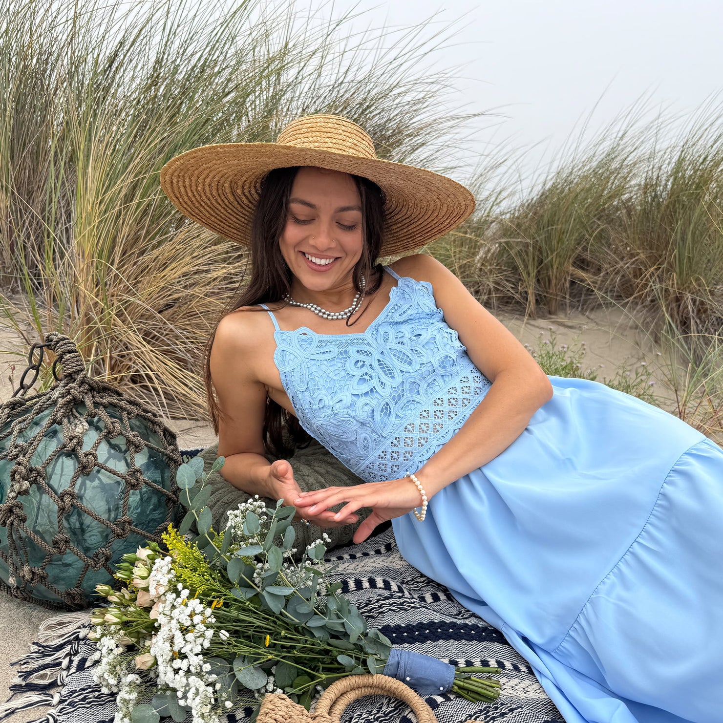 Woman in a blue lace dress and straw hat sitting on a blanket with flowers and a fishing float on a beach wearing white akoya pearl jewerly.