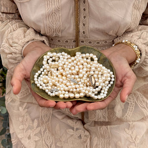 A woman in a tan lace dress is holding a bowl full of lustrous white saltwater akoya pearl necklaces.